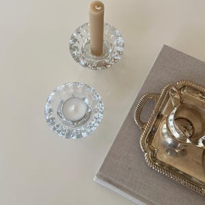 May include: Two clear glass candle holders with a textured, faceted design. One holds a beige candle, the other a white tea light. A silver serving tray and a book are also visible, suggesting a home decor theme.