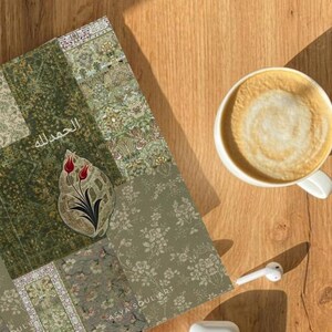 Pu&ograve; includere: Un taccuino con il testo arabo "الحمد لله" e un motivo floreale, accanto a una tazza di caff&egrave; e un paio di auricolari wireless bianchi e una custodia di ricarica. Il taccuino ha un motivo floreale verde.