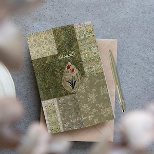 Pu&ograve; includere: Un diario decorativo con un design patchwork nei toni del verde e beige. La copertina presenta un motivo floreale e il testo arabo "الحمد لله". Una penna dorata &egrave; appoggiata accanto al diario su una busta neutra.