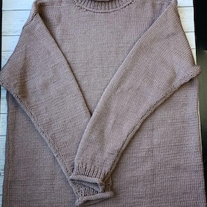 May include: A knitted taupe turtleneck jumper with long sleeves. The jumper has ribbed cuffs and a ribbed turtleneck collar. The sleeves are folded over each other.