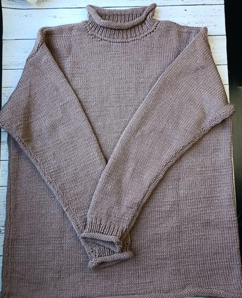 May include: A knitted taupe turtleneck jumper with long sleeves. The jumper has ribbed cuffs and a ribbed turtleneck collar. The sleeves are folded over each other.