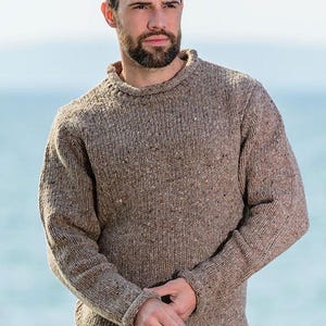 May include: A brown, flecked knitted jumper with a crew neck and ribbed cuffs. The jumper has a textured appearance and a classic, casual style. The model is wearing the jumper outdoors.