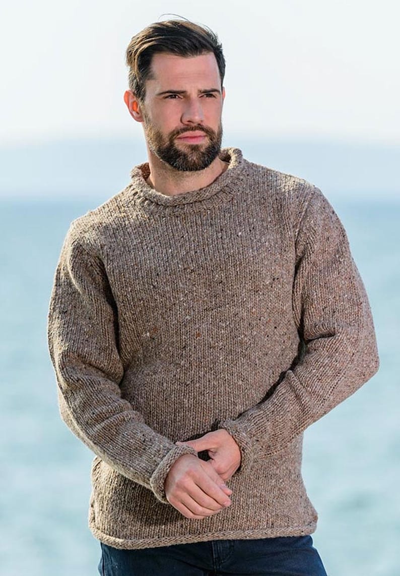 May include: A brown, flecked knitted jumper with a crew neck and ribbed cuffs. The jumper has a textured appearance and a classic, casual style. The model is wearing the jumper outdoors.