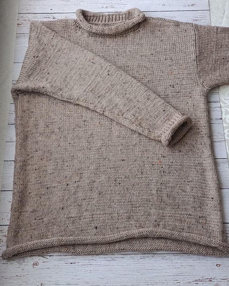 May include: A beige, long-sleeved jumper with a roll neck collar. The jumper has a textured knit pattern with flecks of darker colours throughout. The sleeves are long, and the hemline is straight. The jumper is made of a soft, warm material.