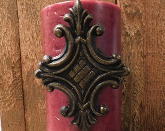 May include: A burgundy pillar candle with a decorative metal accent in the shape of a fleur-de-lis.