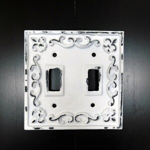 May include: White double light switch cover with an ornate, distressed design. The cover has a white background with black accents.