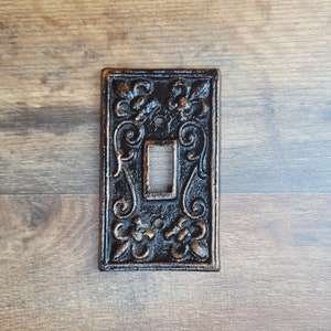 以下が含まれることがあります： 装飾的なデザインが施された黒い鋳鉄製のライトスイッチカバー。カバーにはスイッチ用の開口部が1つあり、標準的なライトスイッチに適合するように設計されています。
