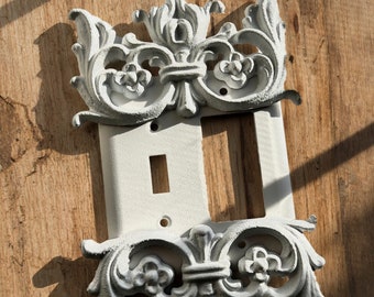May include: White painted metal light switch cover with an ornate, decorative design. The cover is designed to fit a double switch.