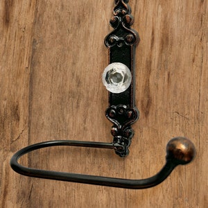 May include: Black metal toilet paper holder with an ornate design and a clear glass knob. The holder is mounted on a wooden wall.