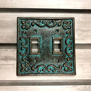 May include: A decorative light switch cover with a turquoise and bronze finish. The cover features a detailed ornate design with scrollwork and fleur-de-lis accents.
