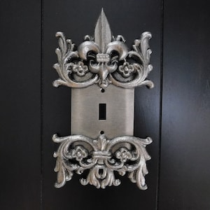 May include: Silver light switch cover with ornate fleur-de-lis design.