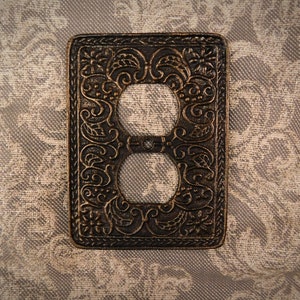Könnte beinhalten: Eine dekorative Steckdosenabdeckung mit einem verzierten, komplizierten Design in einer dunkelbraunen Bronze-Oberfläche.