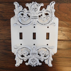 May include: White porcelain light switch cover with a decorative, ornate, white cast iron frame. The frame has a distressed, aged look.