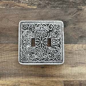 May include: A silver light switch cover with an intricate floral design. The cover has two openings for light switches.