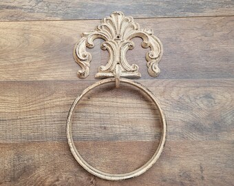 May include: A white metal towel ring with a decorative, ornate, scroll-like design. The ring is mounted on a wooden wall.