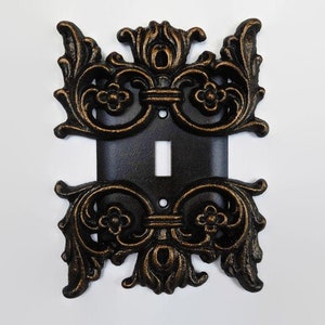 May include: A decorative light switch cover with an ornate, gold-painted, cast iron design. The cover is black and has a single switch opening.