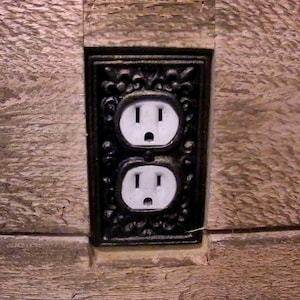 May include: A black double electrical outlet with ornate detailing, mounted on a wood panel.