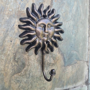 Puede incluir: Un gancho de hierro fundido en forma de sol con un acabado bronce. El gancho tiene una cara con la boca cerrada y los ojos. El gancho está montado en una pared de piedra.