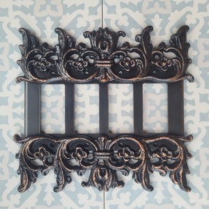 Könnte beinhalten: Eine schwarze Metall-Lichtschalterplatte mit einem verzierten, dekorativen Design. Die Platte hat drei Öffnungen für Lichtschalter.