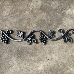 May include: A decorative metal grape vine design with leaves and grape clusters. The metal is a dark silver color. The vine is approximately 12 inches long.