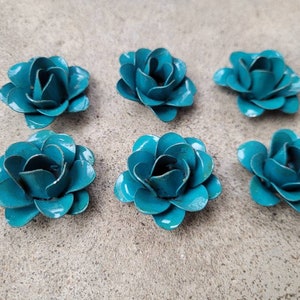May include: Six teal metal rose flower decorations. Each flower has multiple petals and a slightly distressed finish. The flowers are arranged on a neutral-coloured surface.