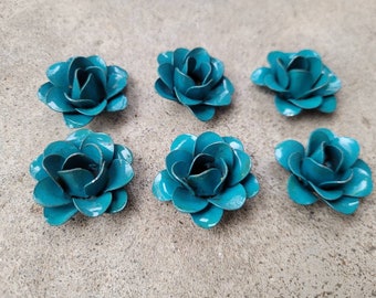 Puede incluir: Seis decoraciones de flores de rosas de metal de color verde azulado. Cada flor tiene múltiples pétalos y un acabado ligeramente desgastado. Las flores están dispuestas sobre una superficie de color neutro.