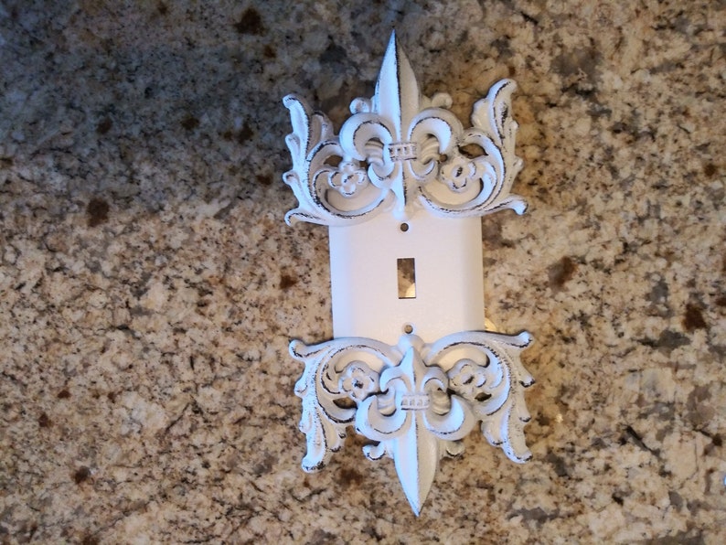May include: White porcelain light switch cover with decorative fleur-de-lis accents.