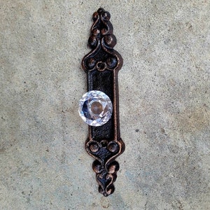 以下が含まれることがあります： 装飾的な、濃いブロンズ色のドアプルで、透明でファセット加工されたクリスタルノブが付いています。プルには装飾的なスクロールワークと長方形の中央があります。ハードウェアは、テクスチャ加工された灰色のコンクリートの表面に配置されています。