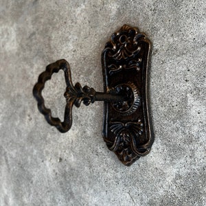 Op de afbeelding: Oud bronzen sleutel en sleutelplaat. De sleutel heeft een decoratieve handgreep en is in het sleutelgat van de plaat gestoken. De plaat heeft een langwerpige vorm met bloemen- en krulwerkdetails. De sleutel en plaat zijn van metaal.
