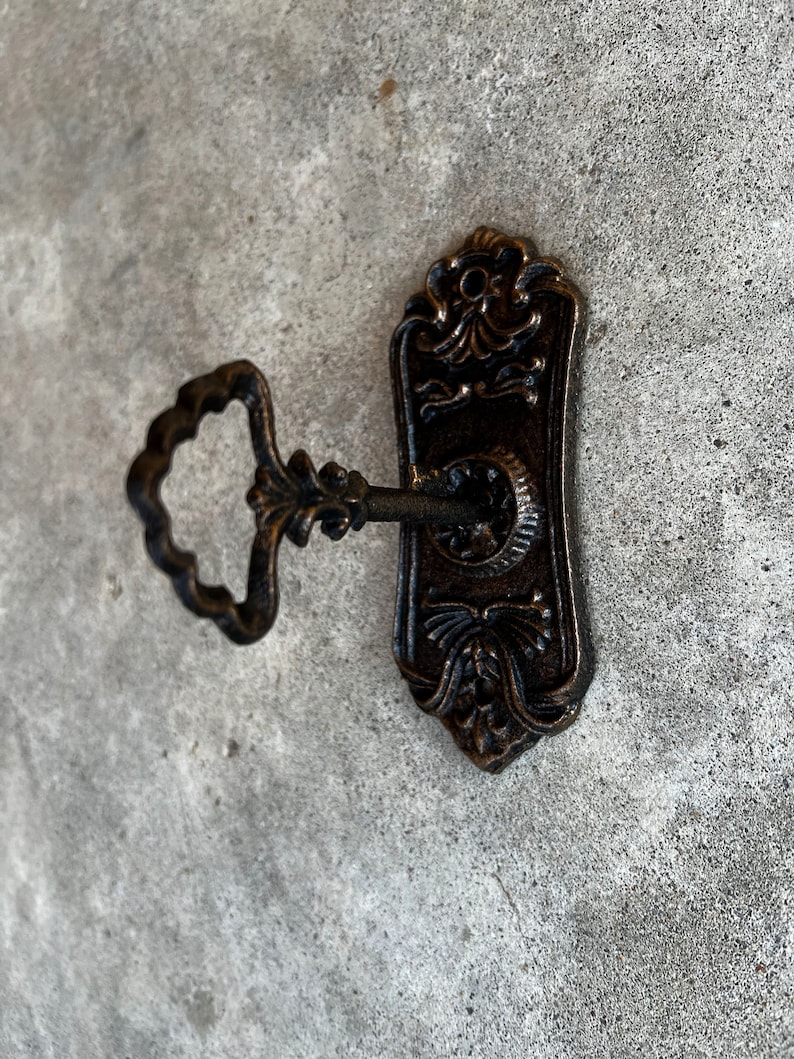 Op de afbeelding: Oud bronzen sleutel en sleutelplaat. De sleutel heeft een decoratieve handgreep en is in het sleutelgat van de plaat gestoken. De plaat heeft een langwerpige vorm met bloemen- en krulwerkdetails. De sleutel en plaat zijn van metaal.