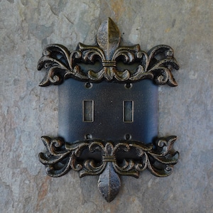 May include: A double light switch plate with a decorative cast iron cover featuring a fleur-de-lis design. The cover is painted in a bronze finish.