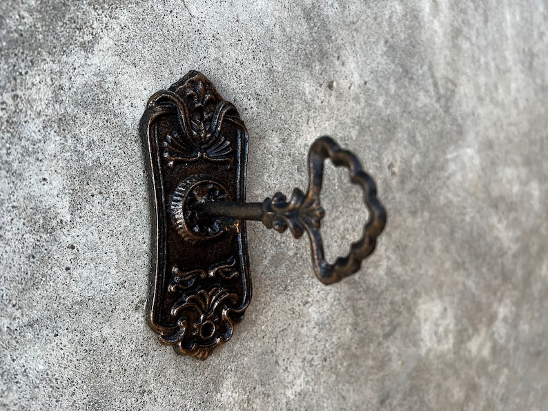 Op de afbeelding: Een sierlijke bronzen sleutel die in een bijpassende sleutelplaat is gestoken. De sleutelplaat heeft een rechthoekige vorm met decoratieve bloemen- en krulwerkdetails. De sleutel heeft een decoratieve handgreep. De achtergrond is een getextureerd grijs oppervlak.