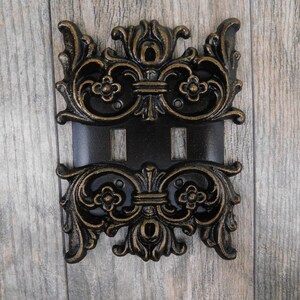 May include: A decorative double light switch cover with an ornate, gold-painted, black metal design.