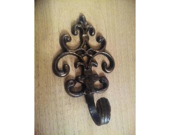 May include: An ornate, dark bronze wall hook with a decorative, scrolled design. The hook has a curved shape and a detailed, textured finish, suitable for hanging items. It is mounted on a light-coloured wooden surface.