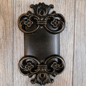 May include: A black cast iron wall plate with an ornate gold finish. The plate has a rectangular shape with a decorative design on the top and bottom. The plate is mounted on a wood surface.