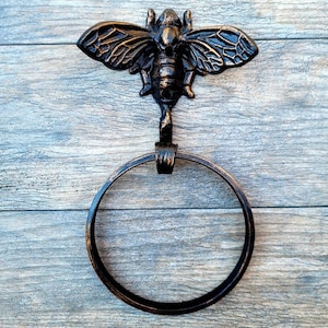 May include: A black metal towel ring with a bee design. The ring is attached to a wooden wall.