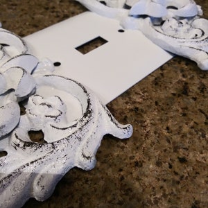 May include: White light switch cover with ornate, white, distressed, metal accents. The accents are in the shape of flowers and leaves.