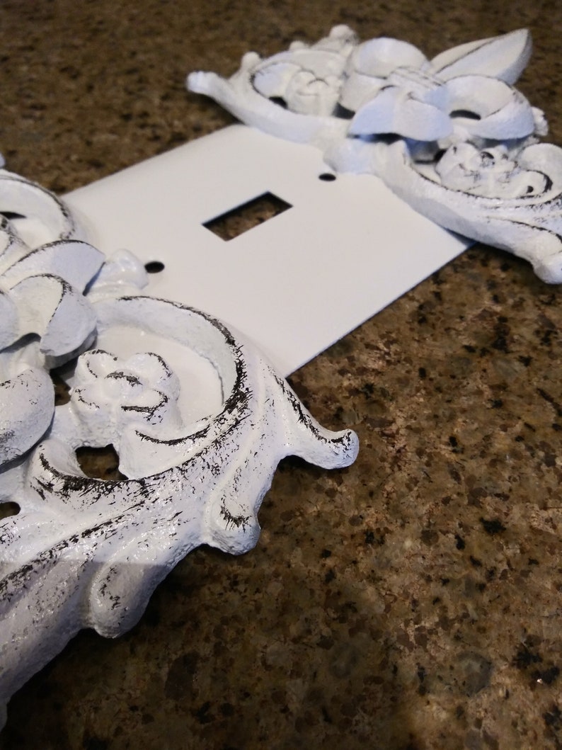 May include: White light switch cover with ornate, white, distressed, metal accents. The accents are in the shape of flowers and leaves.