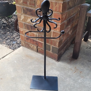 May include: Black metal octopus-themed toilet paper holder. The holder features an octopus design at the top, a circular bar for the roll, and a rectangular base. The holder is freestanding and suitable for a bathroom.