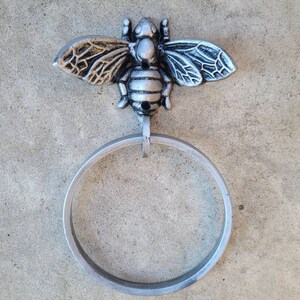 May include: A silver-colored metal towel ring with a decorative bee accent. The bee has detailed wings and a textured body. The ring is circular and hangs below the bee.