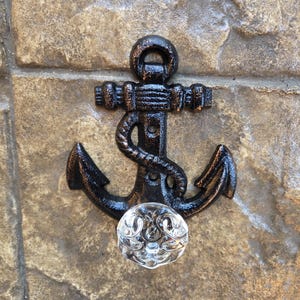 May include: Black anchor-shaped wall hook with a clear, decorative glass knob. The anchor is made of metal and has a rope design on the shank. The hook is mounted on a textured, stone-like surface.