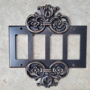 Könnte beinhalten: Eine schwarze Metall-Schalterplatte mit 4 Gangschaltern und verziertem, goldfarbenem Metallbesatz. Der Besatz zeigt ein dekoratives Design mit floralen Motiven.