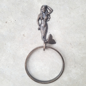 May include: A bronze cast iron towel ring featuring a mermaid design. The mermaid is holding her hair with one hand and has a long flowing tail.