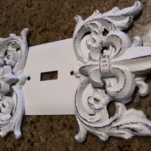 May include: White painted metal light switch cover with decorative fleur-de-lis accents.