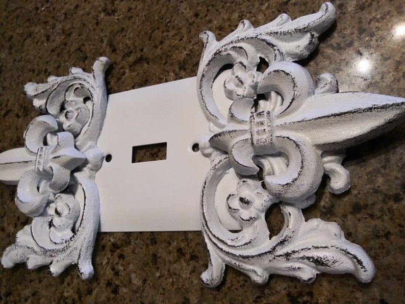 May include: White painted metal light switch cover with decorative fleur-de-lis accents.