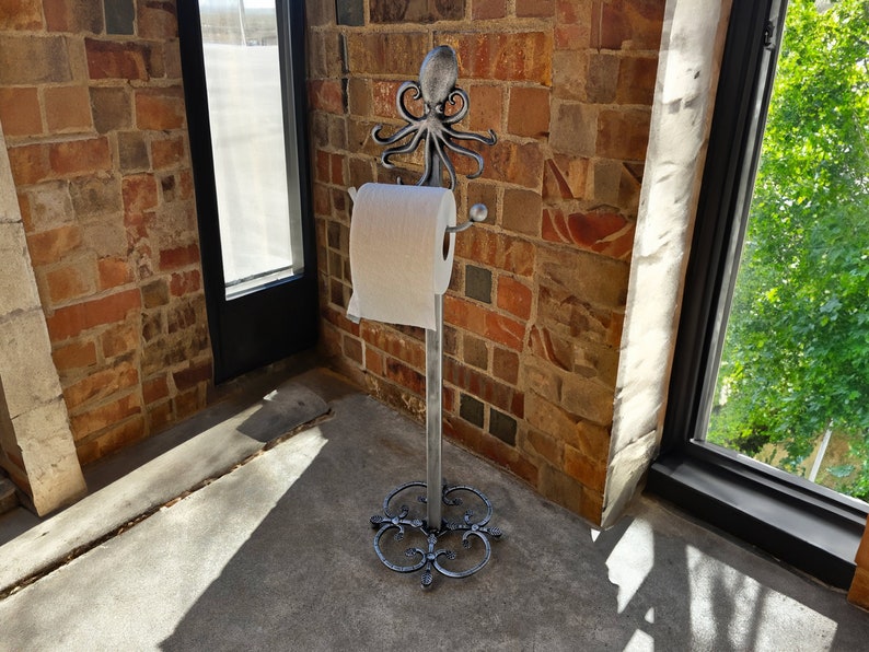 May include: A silver metal toilet roll holder with an octopus design. The holder is standing on a concrete floor in front of a brick wall.