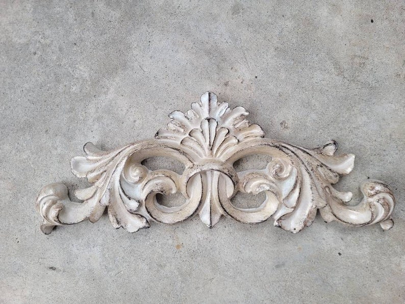 May include: A white, ornate, decorative wall accent with a scroll design. The accent has a distressed, aged look.