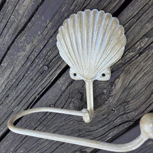 May include: An off-white metal toilet roll holder with a seashell design. The holder features a curved bar for the roll and a decorative shell shape at the top. It is mounted on a weathered wooden surface.