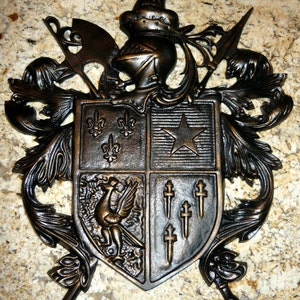 May include: A black metal coat of arms with a shield featuring a star, fleur-de-lis, and a bird. The shield is surrounded by ornate scrollwork and a winged figure.