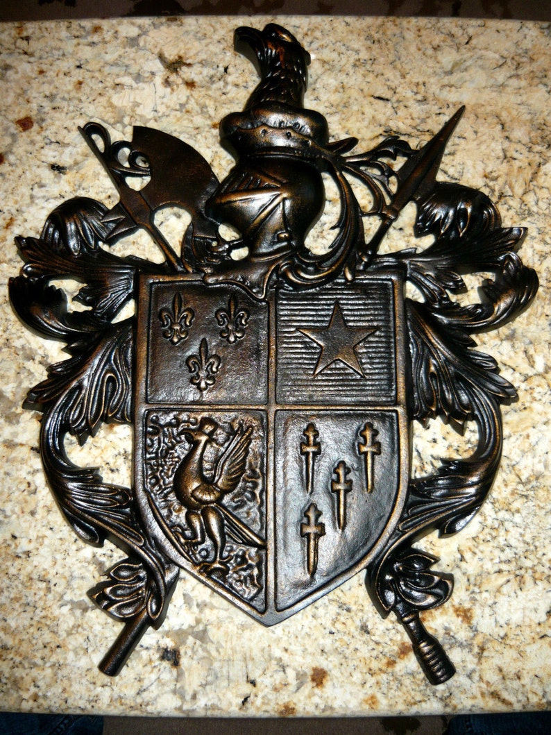 May include: A black metal coat of arms with a shield featuring a star, fleur-de-lis, and a bird. The shield is surrounded by ornate scrollwork and a winged figure.